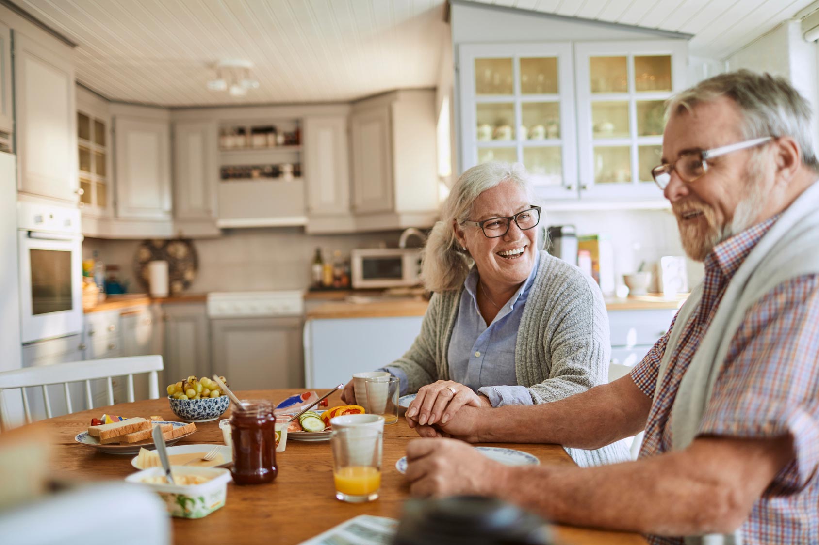 A senior-aged man and woman smiling, sitting at a kitchen table - Carolina Heating Service Inc. A senior-aged man and woman smiling, sitting at a kitchen table